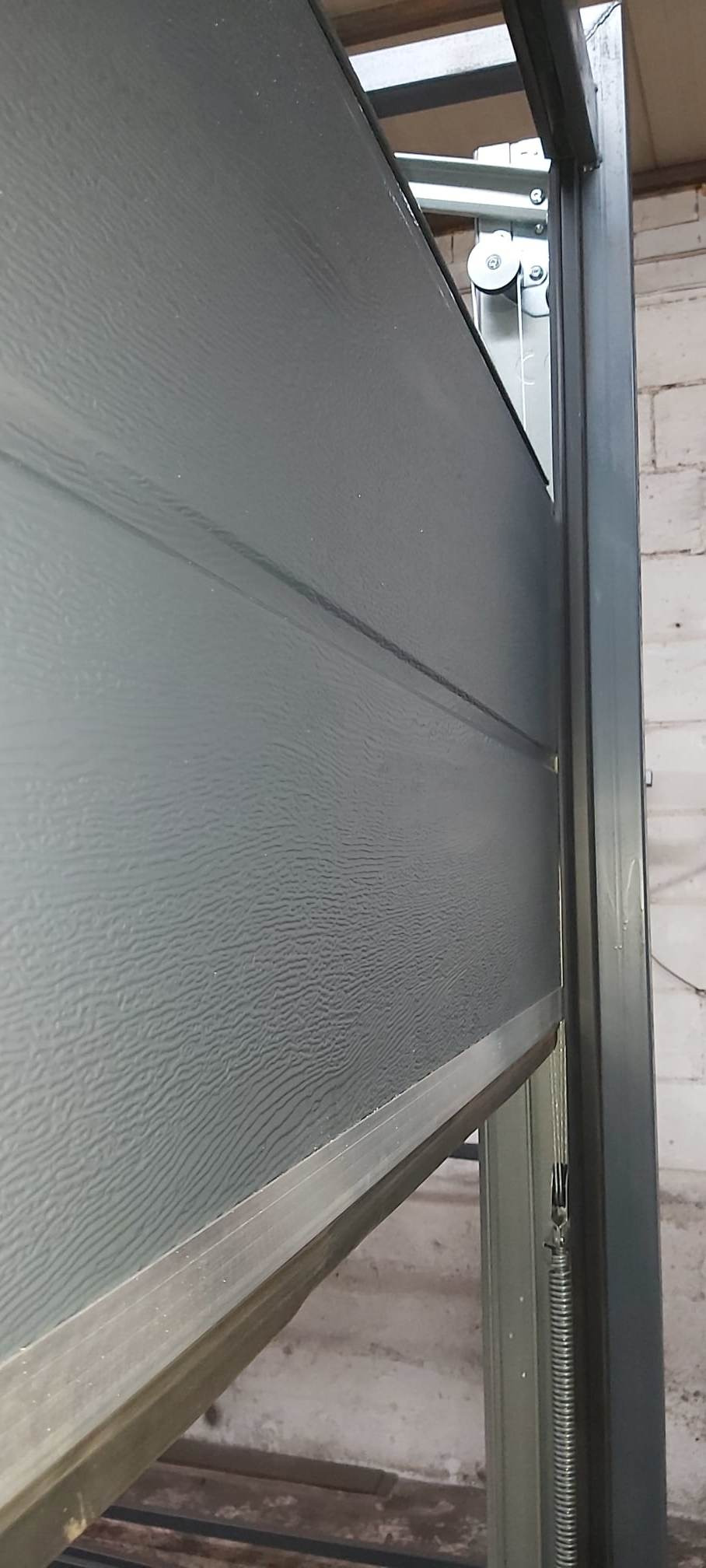 Fragment szarej bramy segmentowej garażowej z widocznym mechanizmem podnoszenia na tle ściany z cegły.