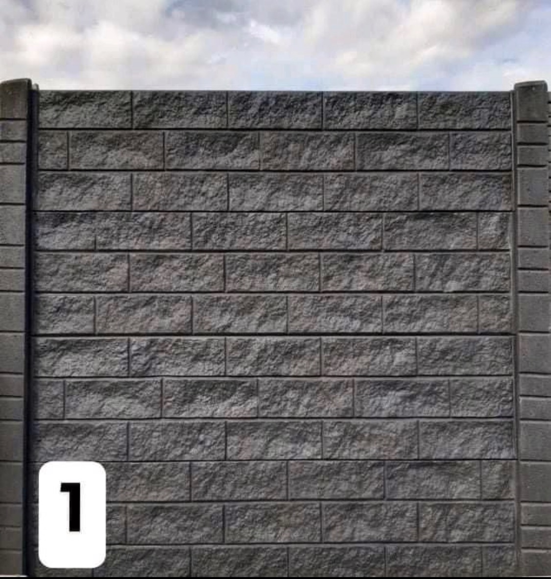 Szary mur z betonowych bloczków imitujących kamień, tworzący fragment ogrodzenia z pionowymi słupkami po bokach, na tle pochmurnego nieba.