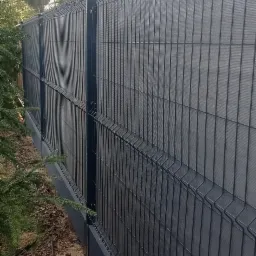 Nowoczesne ogrodzenie panelowe z podmurówką w kolorze antracytowym, zapewniające prywatność dzięki wypełnieniu z siatki maskującej, widoczne z perspektywy bocznej, z fragmentem budynku w tle.