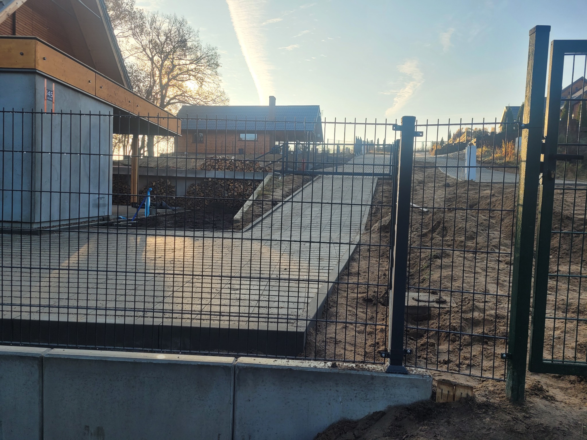 Nowoczesne ogrodzenie panelowe z betonową podmurówką na posesji z domem w tle, widoczne otwarte skrzydło furtki.