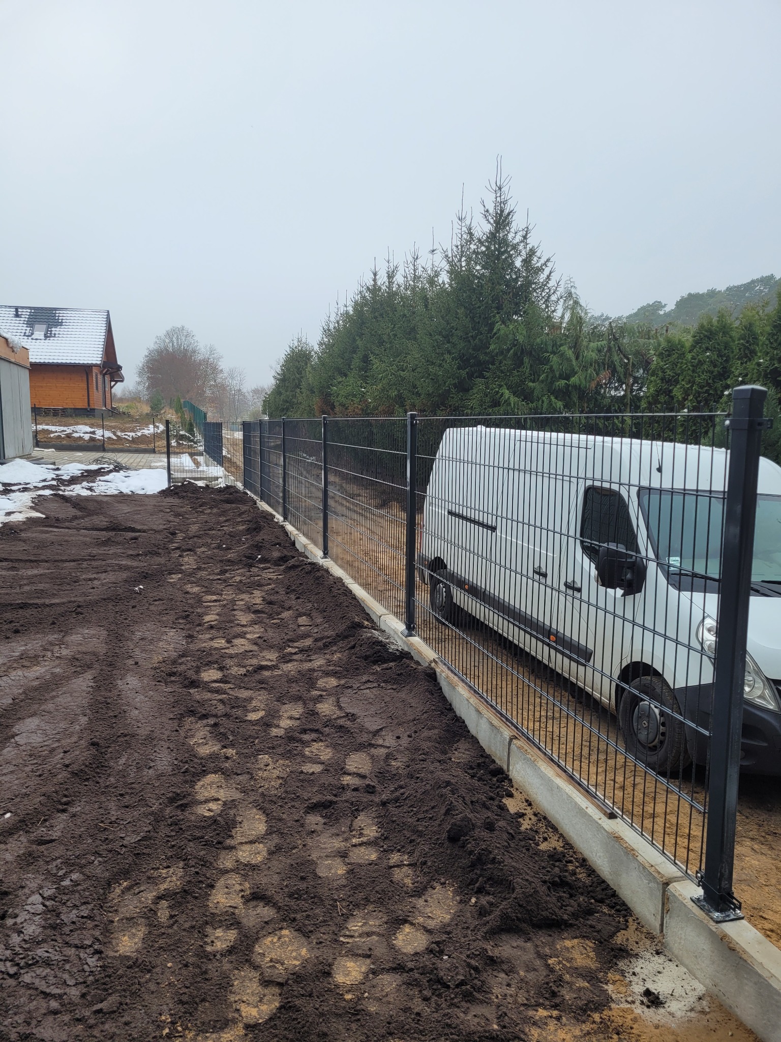 Nowo postawione, czarne, metalowe ogrodzenie panelowe na betonowej podmurówce, z widocznym białym samochodem dostawczym w tle, na błotnistym terenie w pochmurny dzień.
