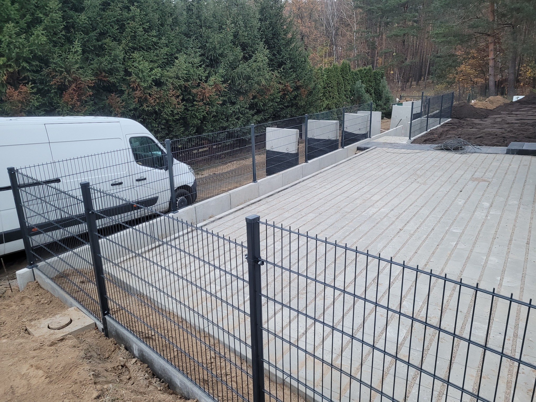 Nowoczesne ogrodzenie panelowe z podmurówką betonową w trakcie montażu, widoczny biały bus dostawczy na tle zielonych drzew.