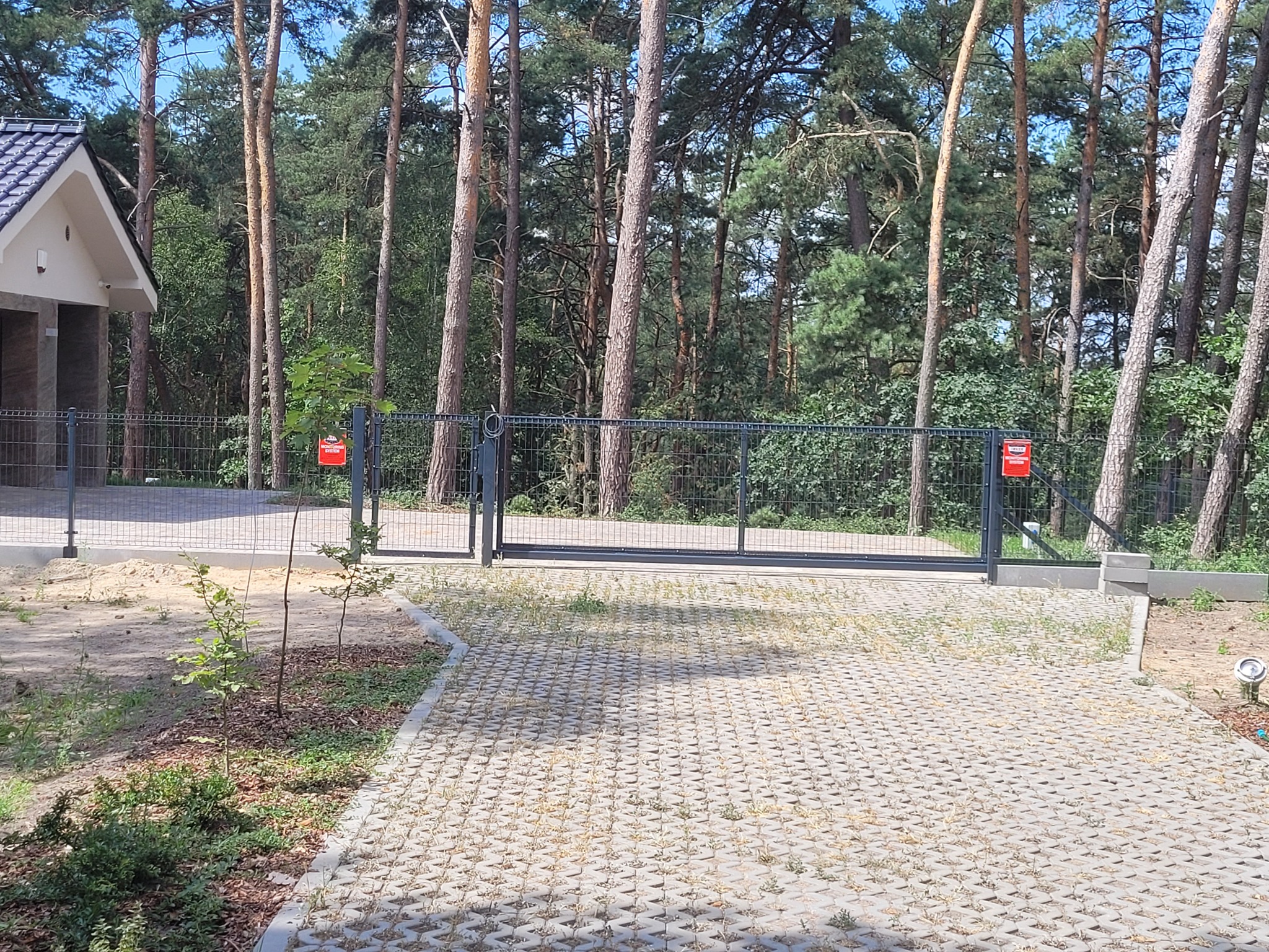 Automatyczna brama przesuwna z metalową siatką ogrodzeniową, widoczna z zewnątrz posesji, z lasem w tle.