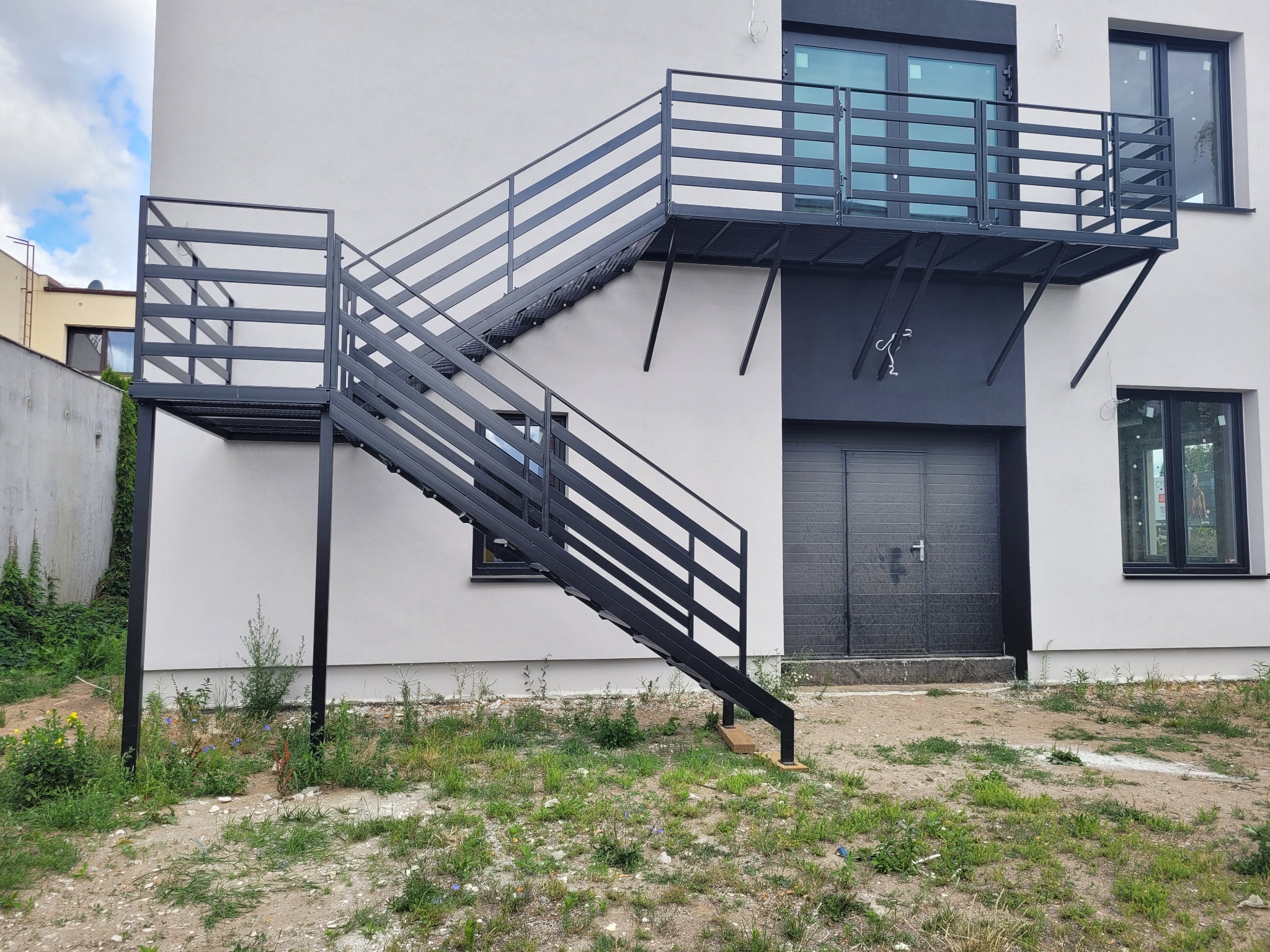 Zewnętrzne, metalowe schody z platformą prowadzące na balkon, przymocowane do ściany budynku z szarą bramą garażową.