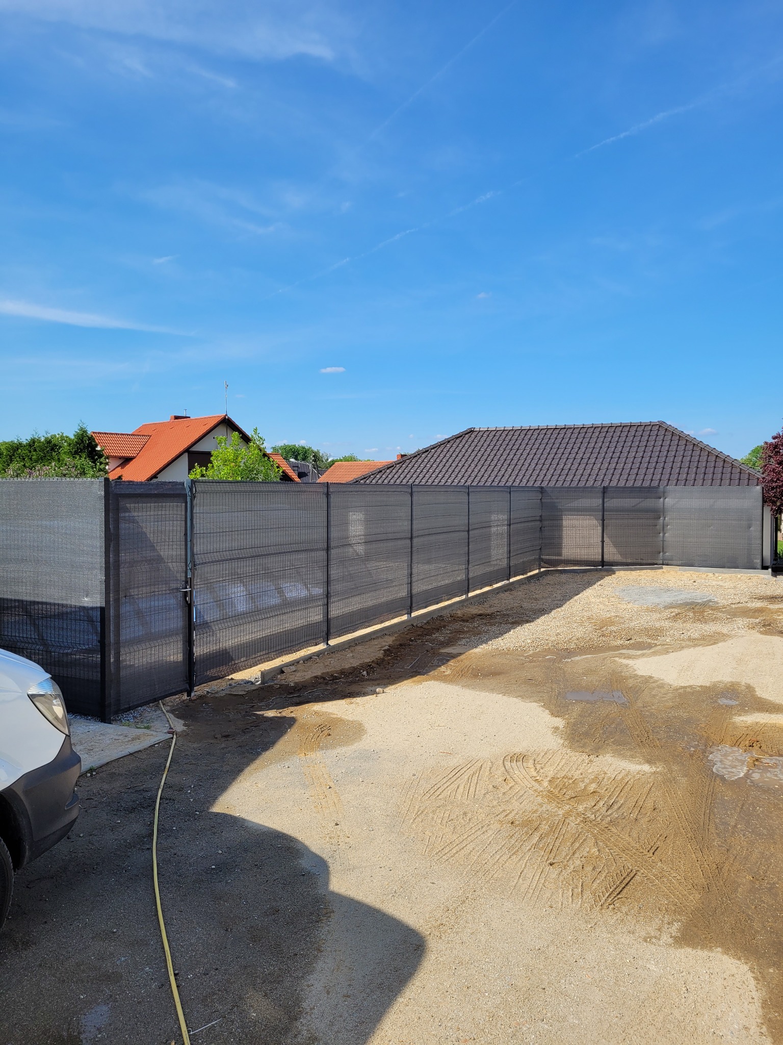 Nowoczesne ogrodzenie panelowe z siatką maskującą w kolorze grafitowym, widok z zewnątrz posesji, częściowo utwardzony teren, błękitne niebo.