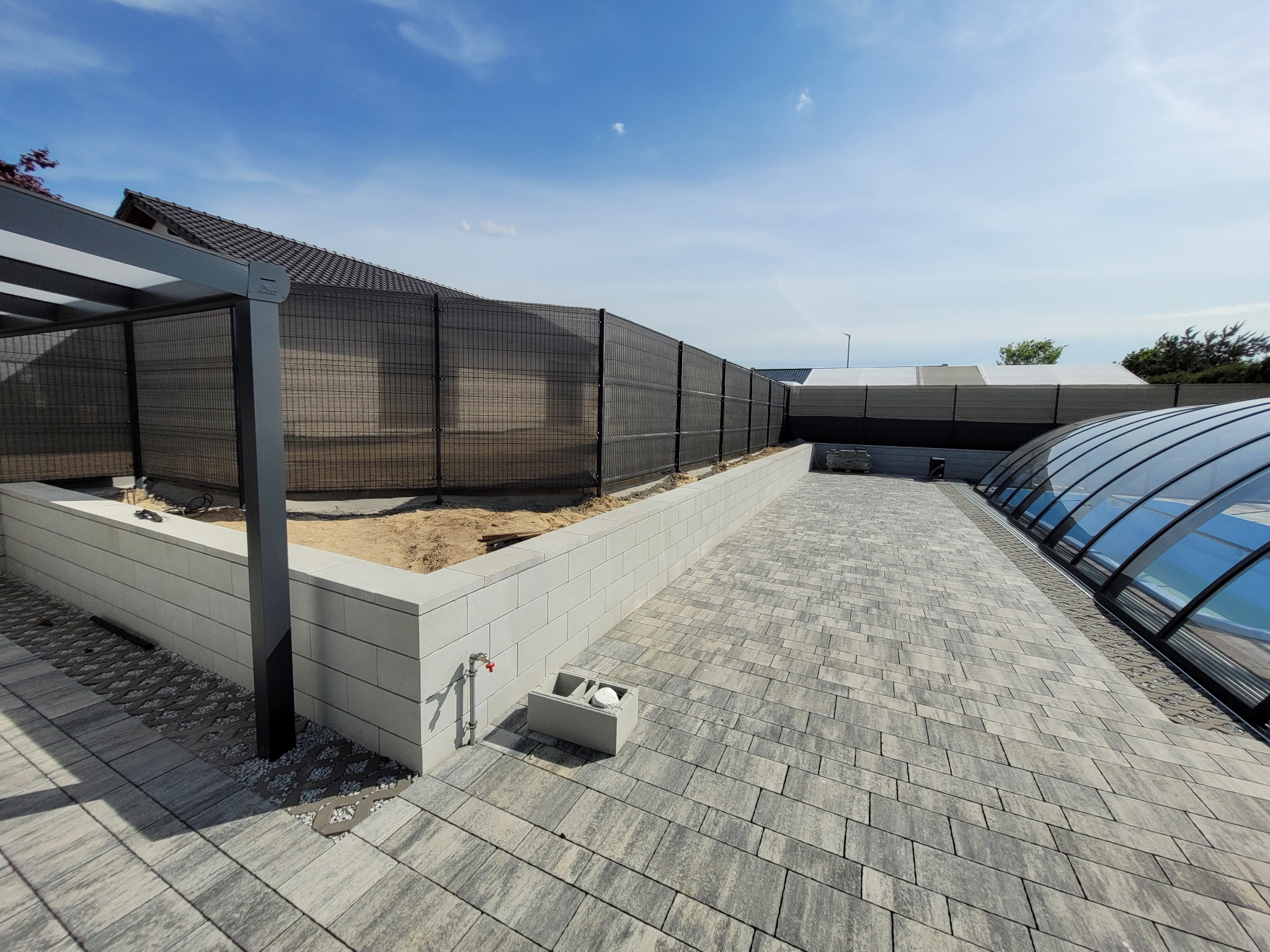 Nowoczesne ogrodzenie panelowe z czarną siatką na podmurówce z szarej kostki betonowej, widok na basen z zadaszeniem i fragment pergoli