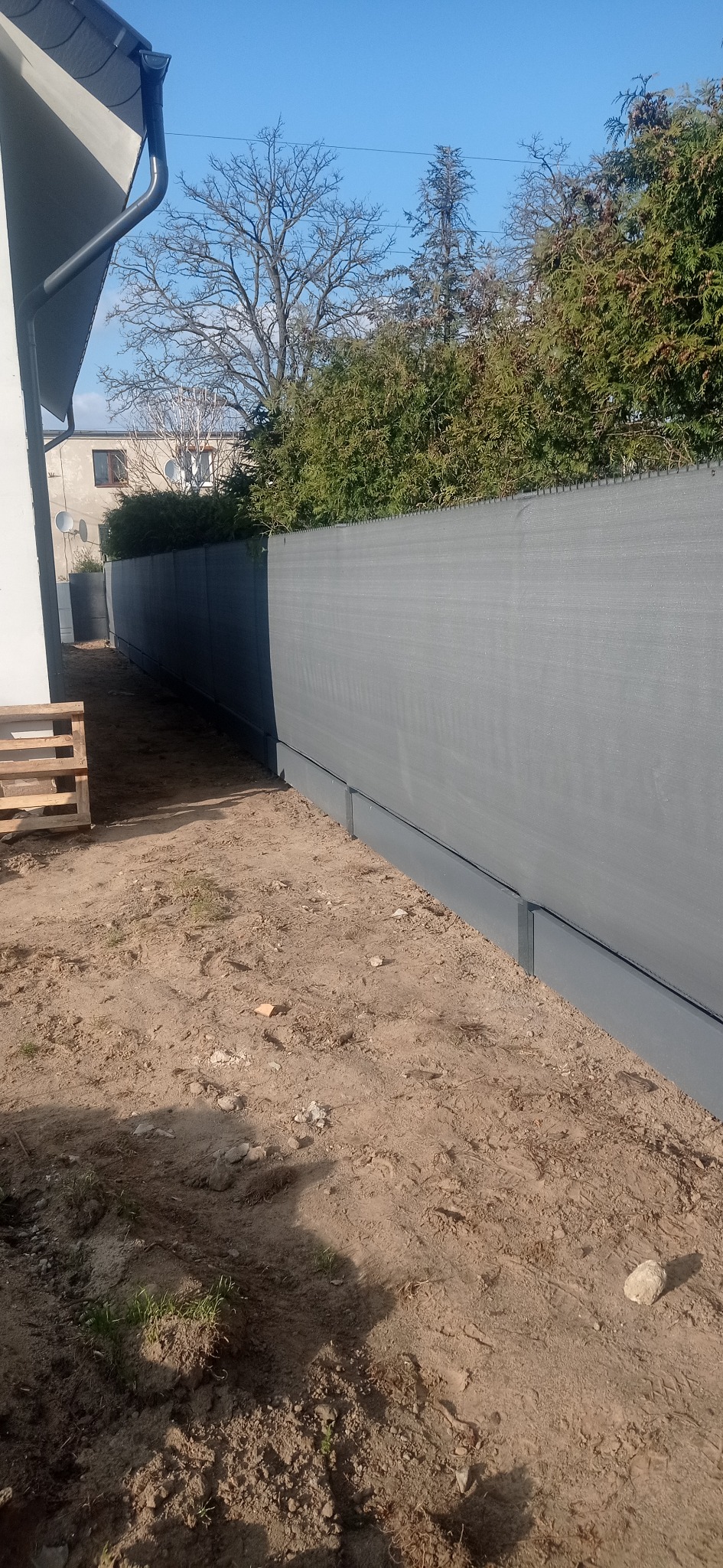 Długi, szary panelowy płot z podmurówką na nierównym, piaszczystym terenie, widok z boku z drewnianą paletą w tle, błękitne niebo w górnej części kadru.