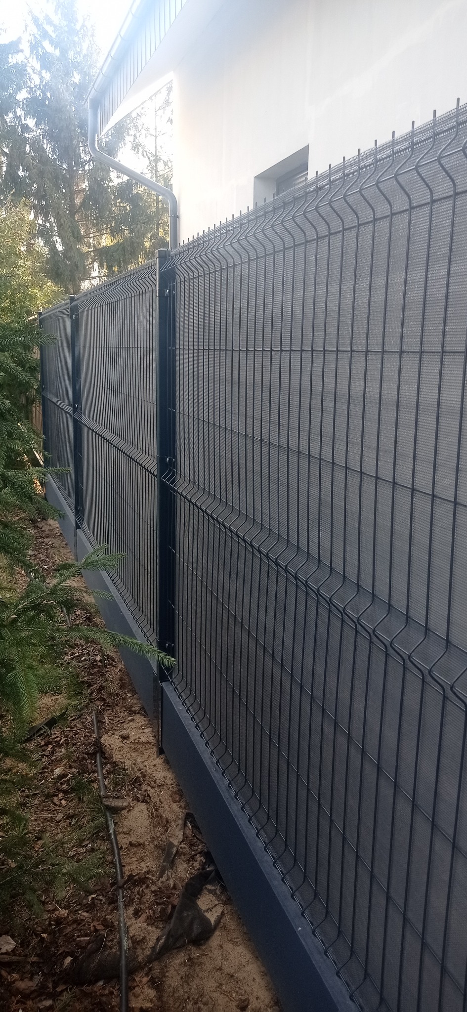 Nowoczesne ogrodzenie panelowe z podmurówką w kolorze antracytowym, zapewniające prywatność dzięki wypełnieniu z siatki maskującej, widoczne z perspektywy bocznej, z fragmentem budynku w tle.