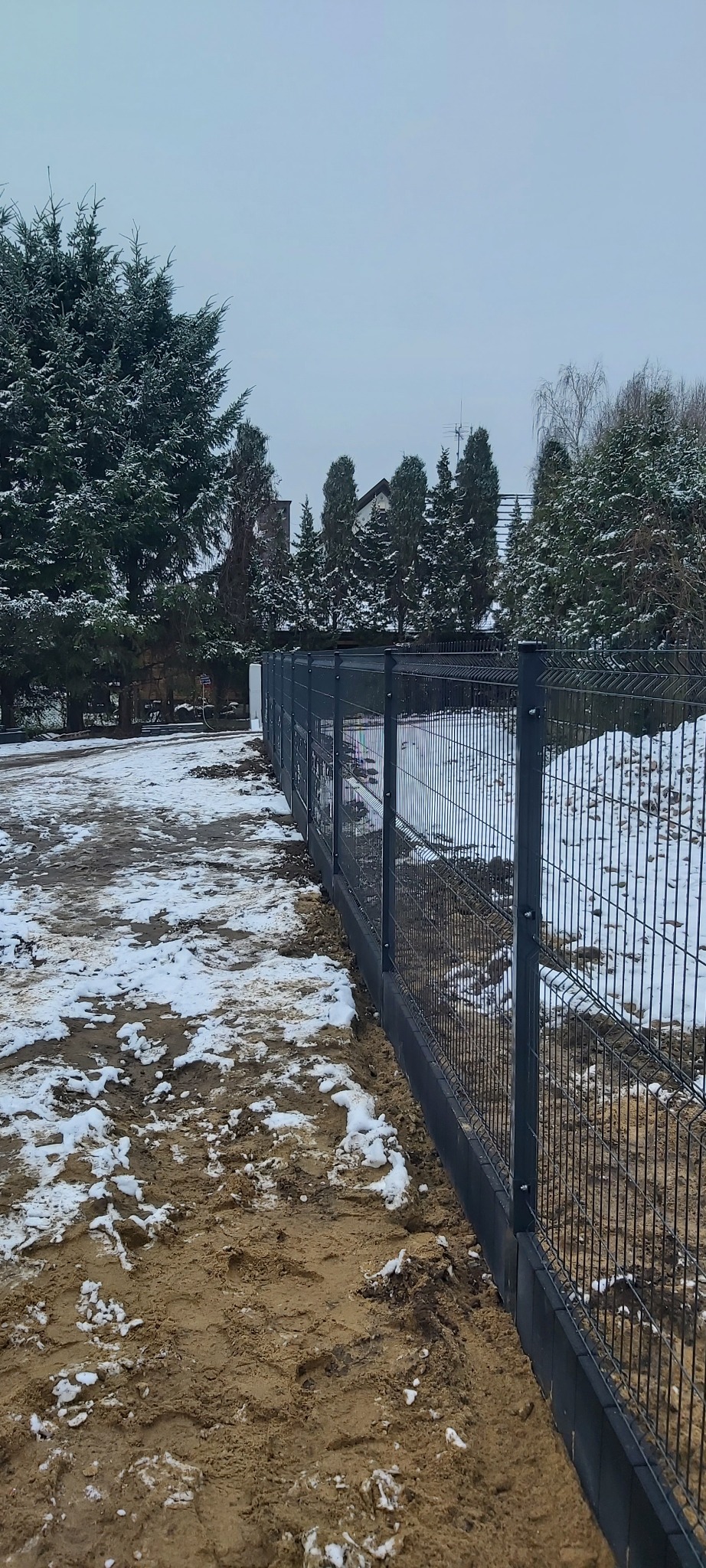 Nowo postawione, ciemnoszare ogrodzenie panelowe z podmurówką betonową, widoczne na tle zasypanego śniegiem terenu i drzew w pochmurny dzień.