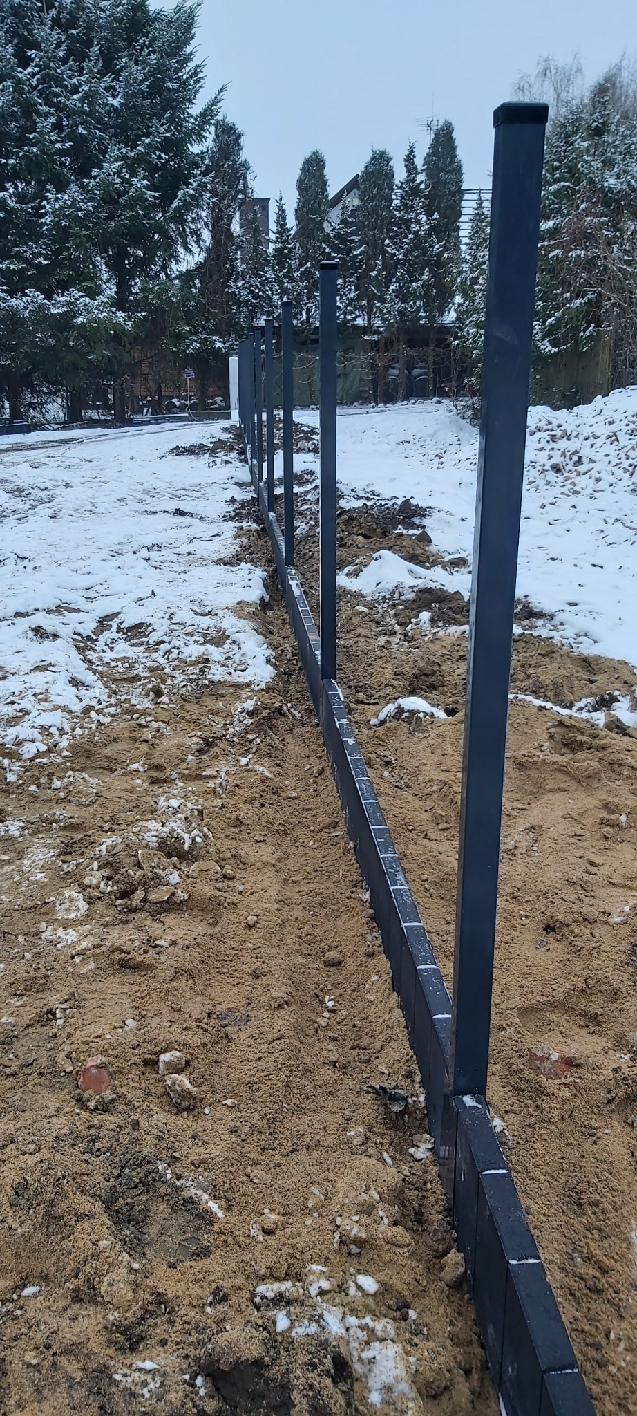 Wykop pod fundament ogrodzenia z metalowymi słupkami w Wyszkowie, widoczne ślady śniegu na piasku i ziemi, w tle zieleń drzew.