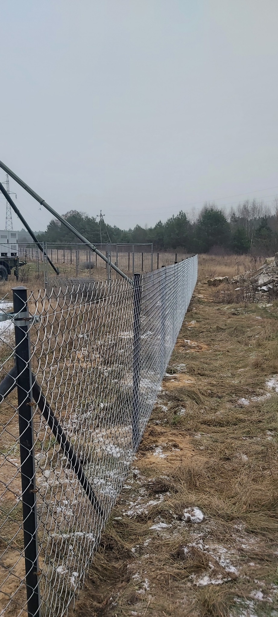 Długi fragment ogrodzenia z siatki, z czarnymi słupkami, ciągnący się wzdłuż pola z suchą trawą i resztkami śniegu, na tle lasu i pochmurnego nieba.