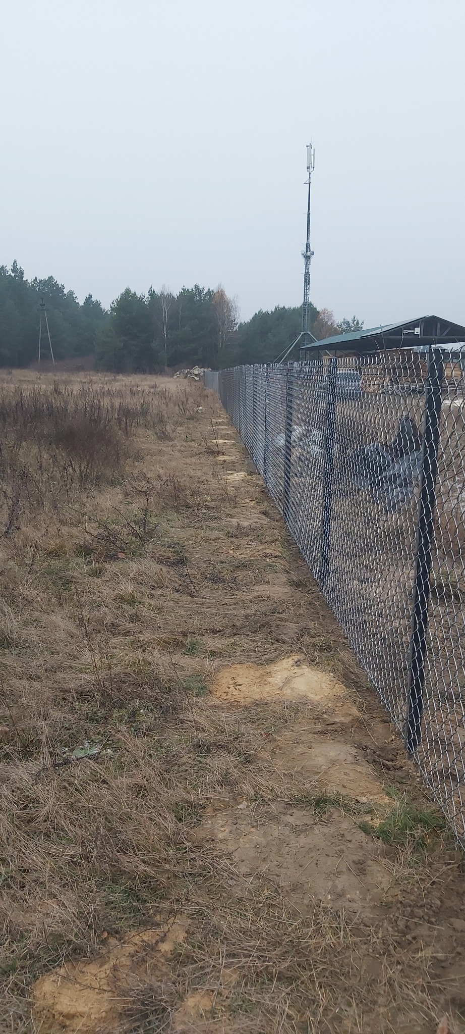 Długi, prosty odcinek ogrodzenia z siatki leśnej rozciągający się wzdłuż polnej drogi, z widoczną wieżą telekomunikacyjną w tle i stertą kamieni po lewej stronie