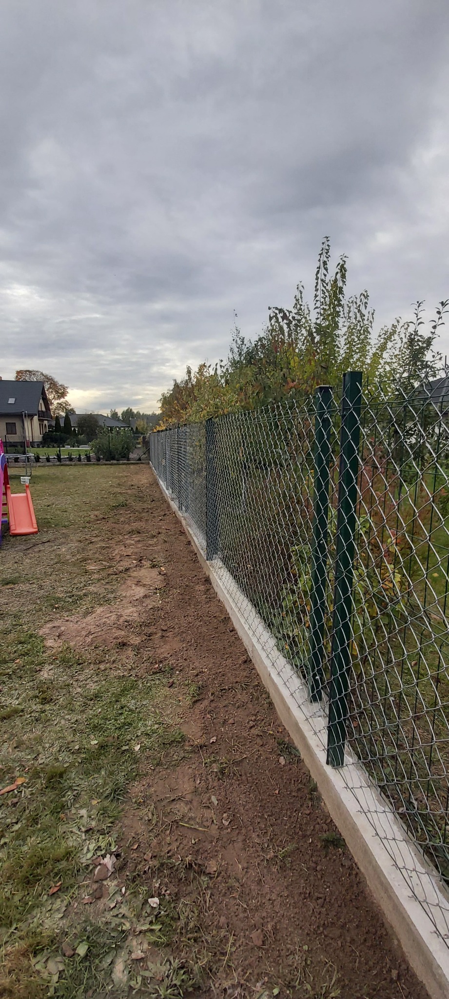 Długa siatka ogrodzeniowa z betonową podmurówką, widok z boku, za ogrodzeniem drzewa i dom jednorodzinny, pochmurne niebo.