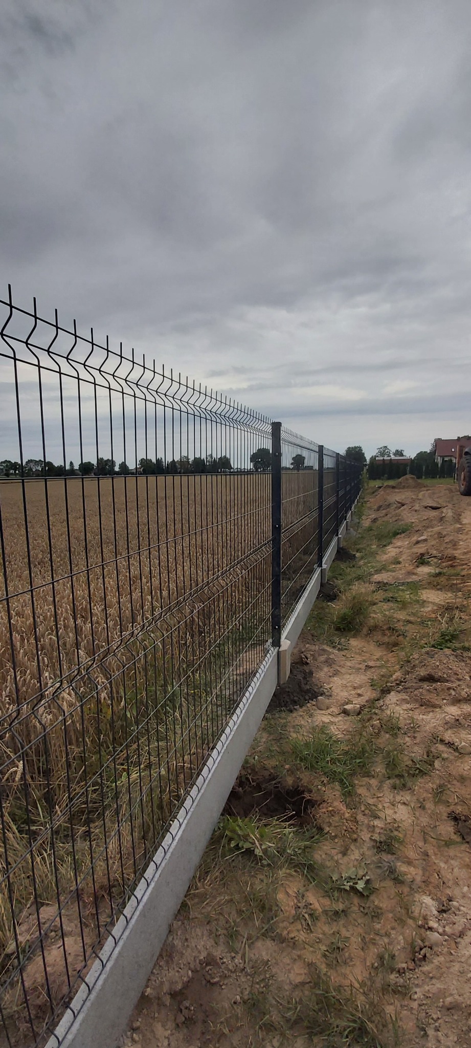 Długi, czarny panelowy system ogrodzeniowy z podmurówką betonową, oddzielający pole zboża od trawnika, z widocznym traktorem w tle i pochmurnym niebem