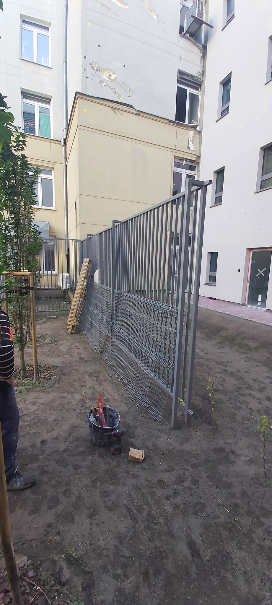 Fragment montażu ogrodzenia panelowego z widocznym wiadrem z narzędziami, wiertarką i ułożonymi panelami ogrodzeniowymi na tle budynków.