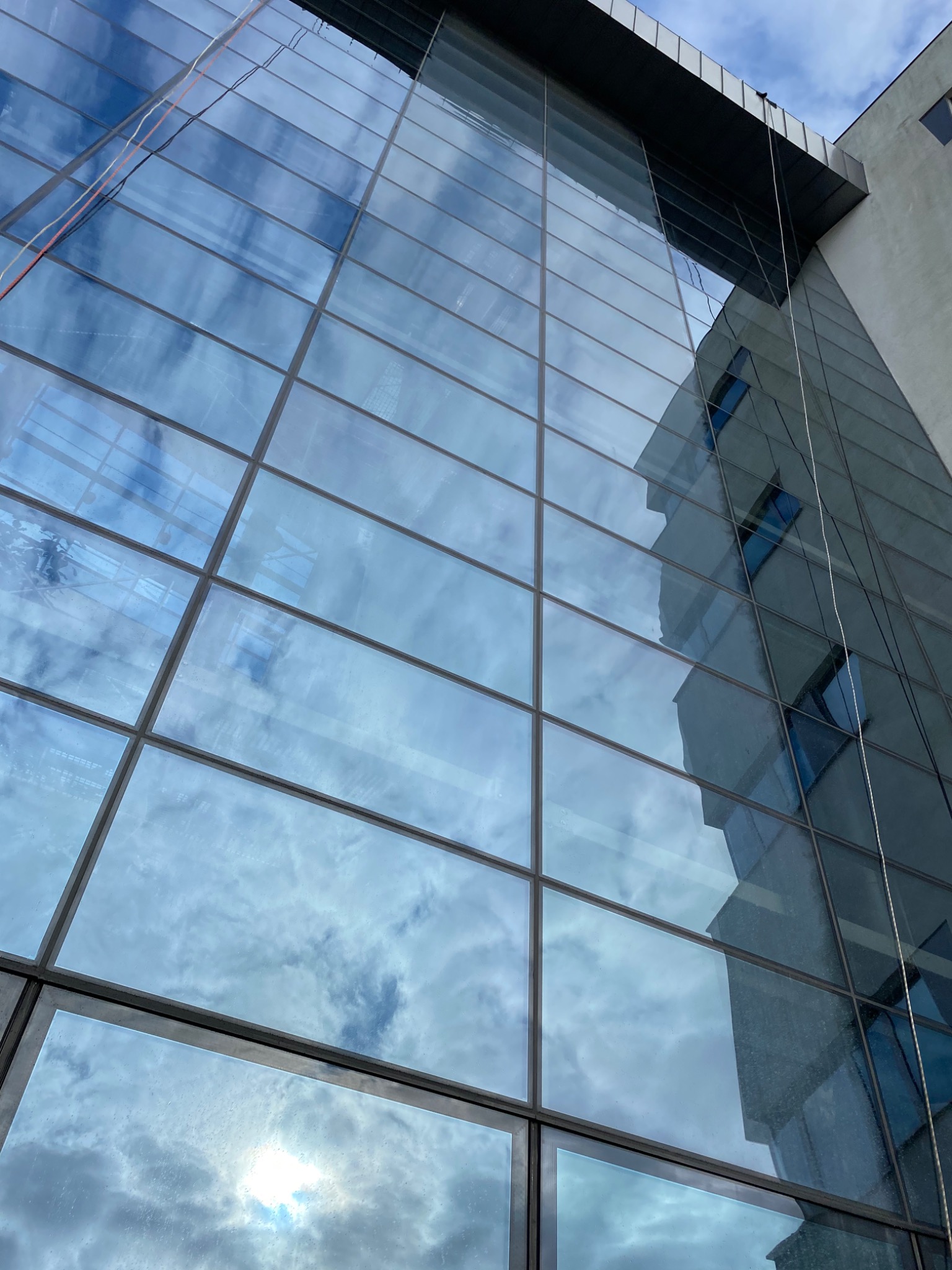 Szklana fasada budynku biurowego z widocznymi odbiciami chmur i sąsiedniego budynku, z liną do mycia okien zwisającą z góry.