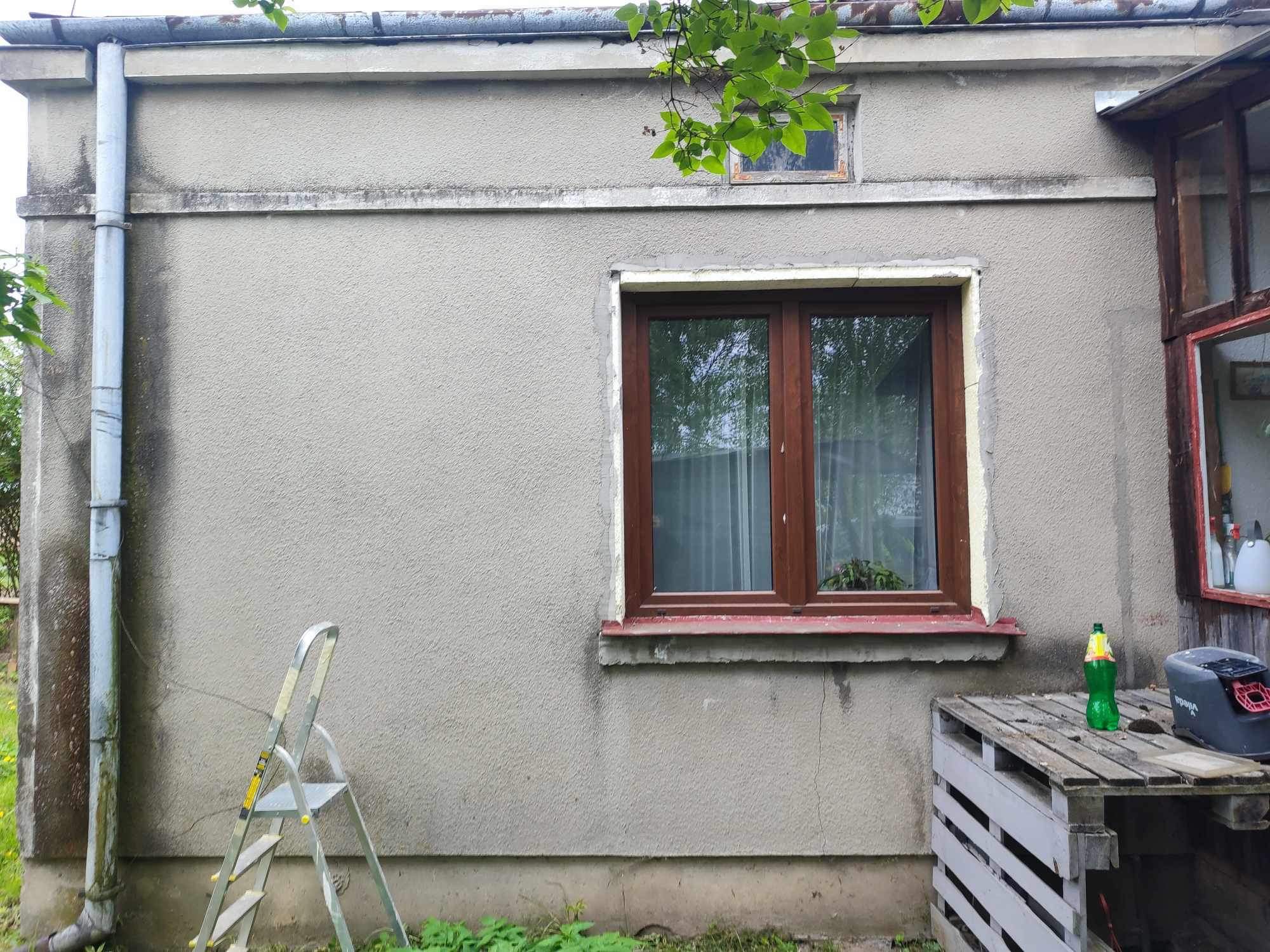 Szara elewacja budynku z drewnianym oknem, drabina oparta o ścianę, widoczny fragment rynny, na palecie butelka i urządzenie Vileda.