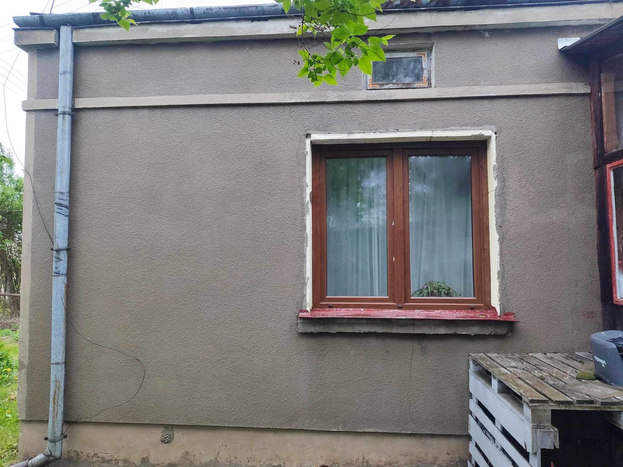 Szara elewacja budynku z drewnianą ramą okienną, widoczne ślady napraw i malowania, rynna z boku budynku, drewniana paleta pod oknem, gałązka drzewa w górnym rogu.