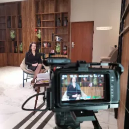 Kamera ustawiona na statywie nagrywa kobietę siedzącą na krześle w eleganckim wnętrzu z drewnianymi regałami w tle. Na monitorze kamery widoczny podgląd nagrywanej sceny.