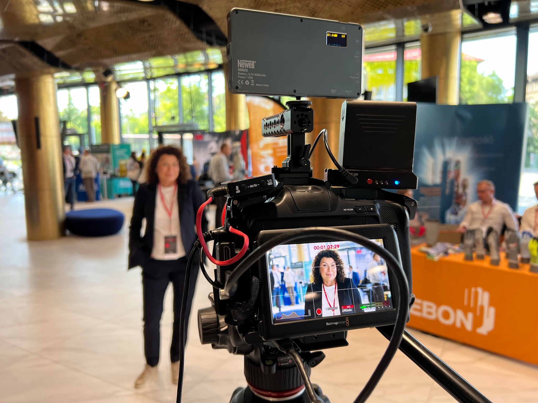 Profesjonalna kamera Blackmagic Design z podglądem nagrywanej osoby podczas konferencji, oświetlenie Newell Rangha, widoczne okablowanie i statyw, w tle uczestnicy wydarzenia.