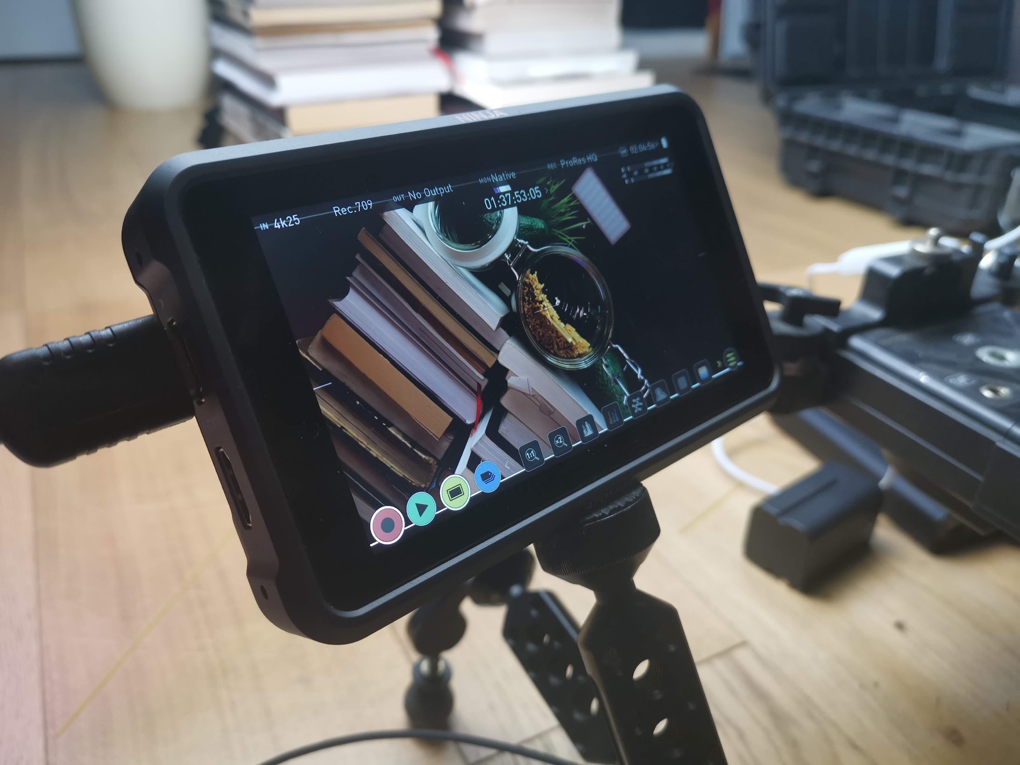 Monitor podglądowy na statywie wyświetlający ujęcie książek i słoika z widocznym interfejsem nagrywania, obok widoczny akumulator i inne elementy sprzętu filmowego.