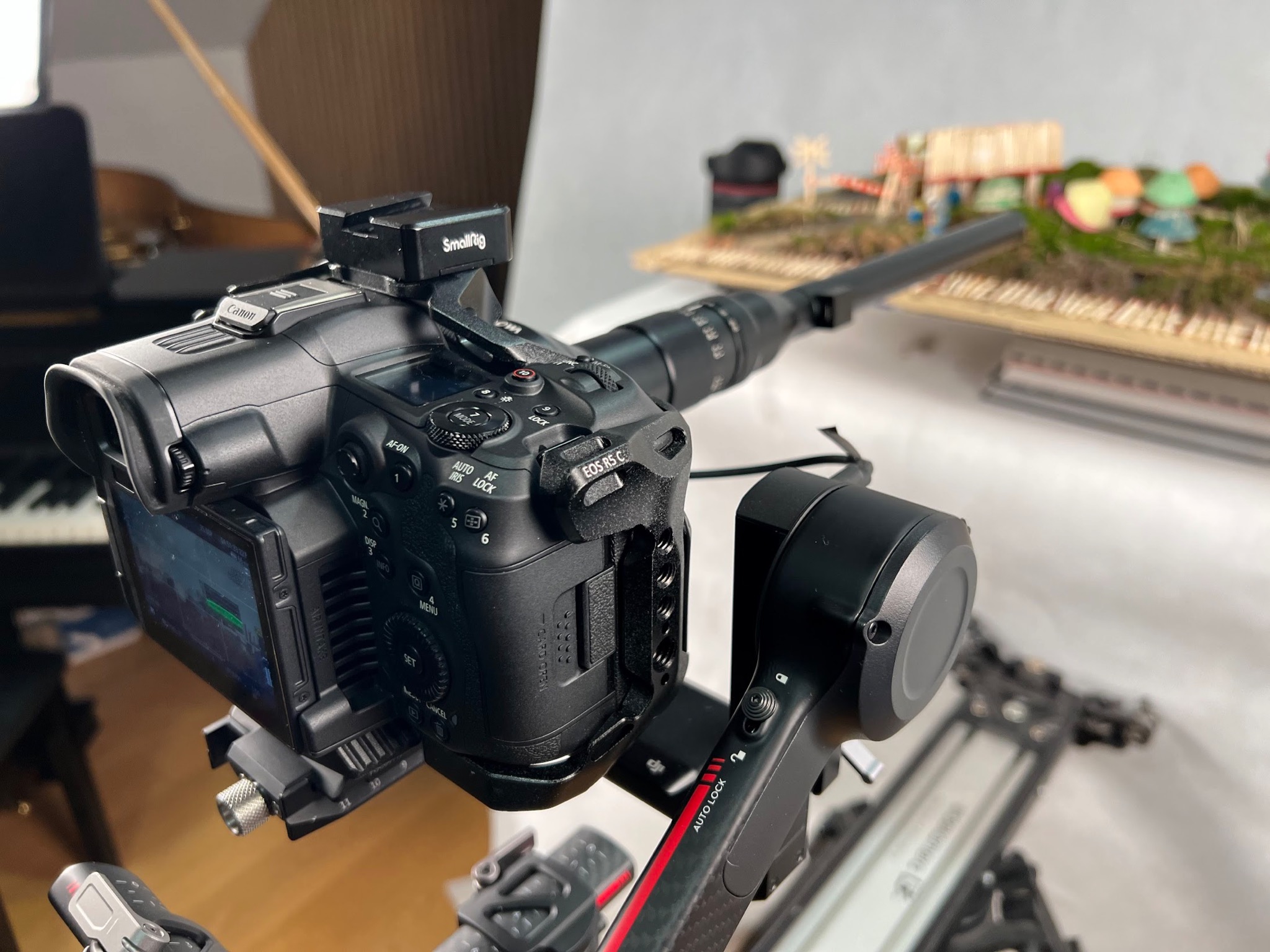 Profesjonalna kamera Canon EOS R5 C zamontowana na gimbalu, ustawiona do filmowania miniaturowej sceny z domkami i drzewami, widoczny ekran LCD z podglądem nagrywanego materiału.