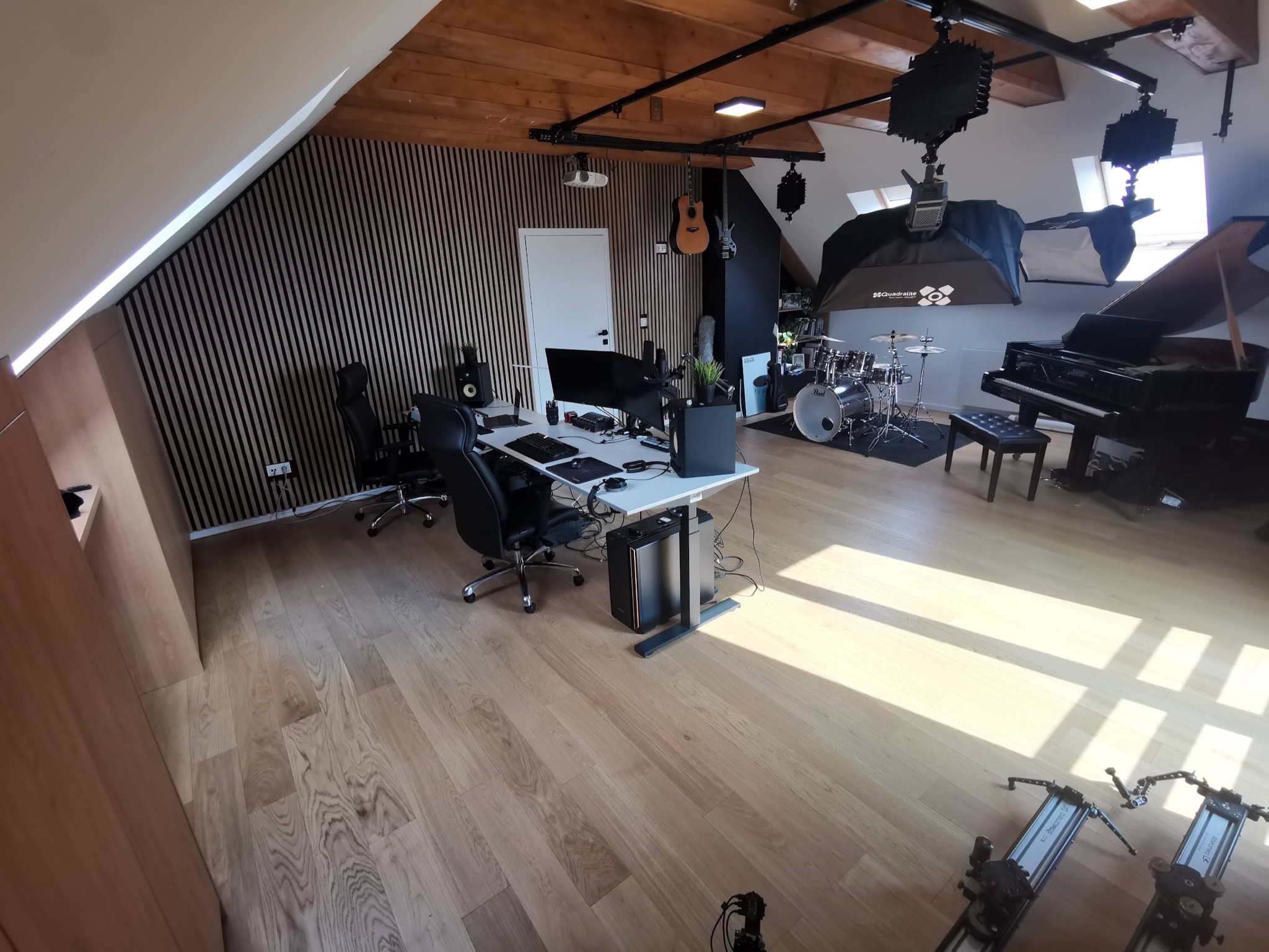 Przestronne studio nagraniowe na poddaszu z instrumentami: pianino, perkusja, gitary, stanowiska komputerowe z monitorami, oświetlenie studyjne na szynach, drewniana podłoga i ściana z paneli...