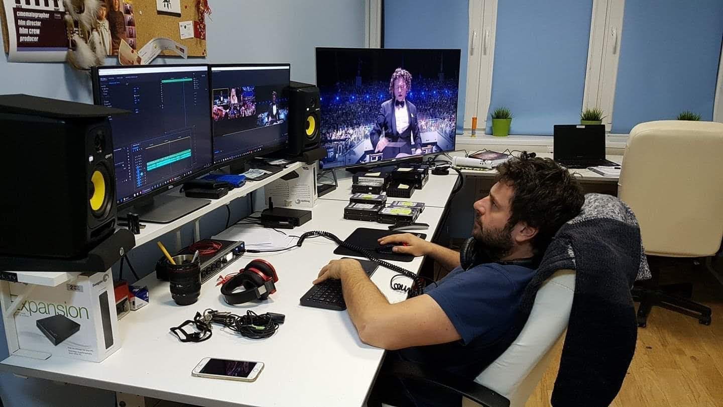 Mężczyzna siedzi przy biurku z dwoma monitorami wyświetlającymi oprogramowanie do edycji wideo, dodatkowy monitor pokazuje występ artysty na scenie, widoczne dyski zewnętrzne, słuchawki i inne...