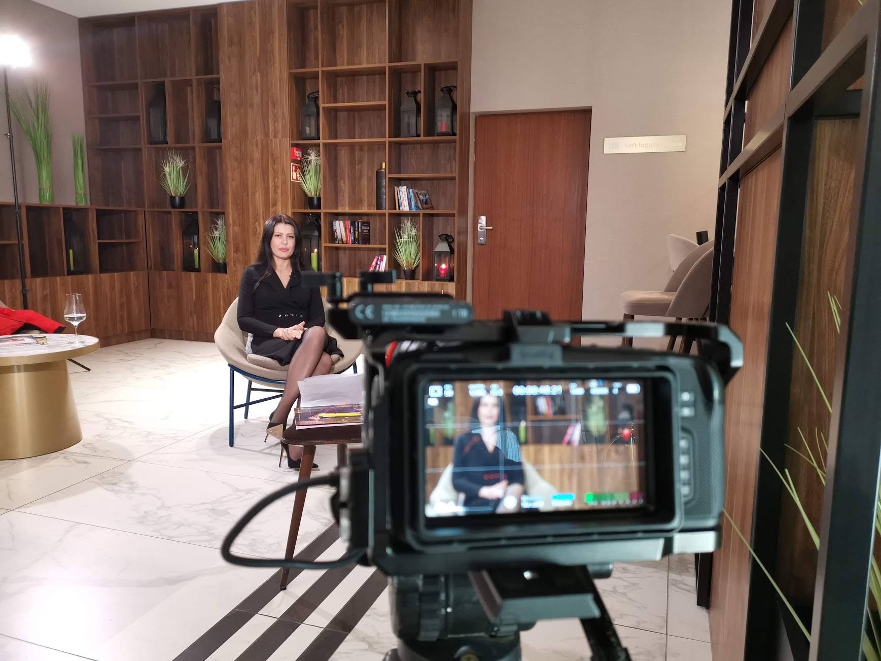 Kamera ustawiona na statywie nagrywa kobietę siedzącą na krześle w eleganckim wnętrzu z drewnianymi regałami w tle. Na monitorze kamery widoczny podgląd nagrywanej sceny.