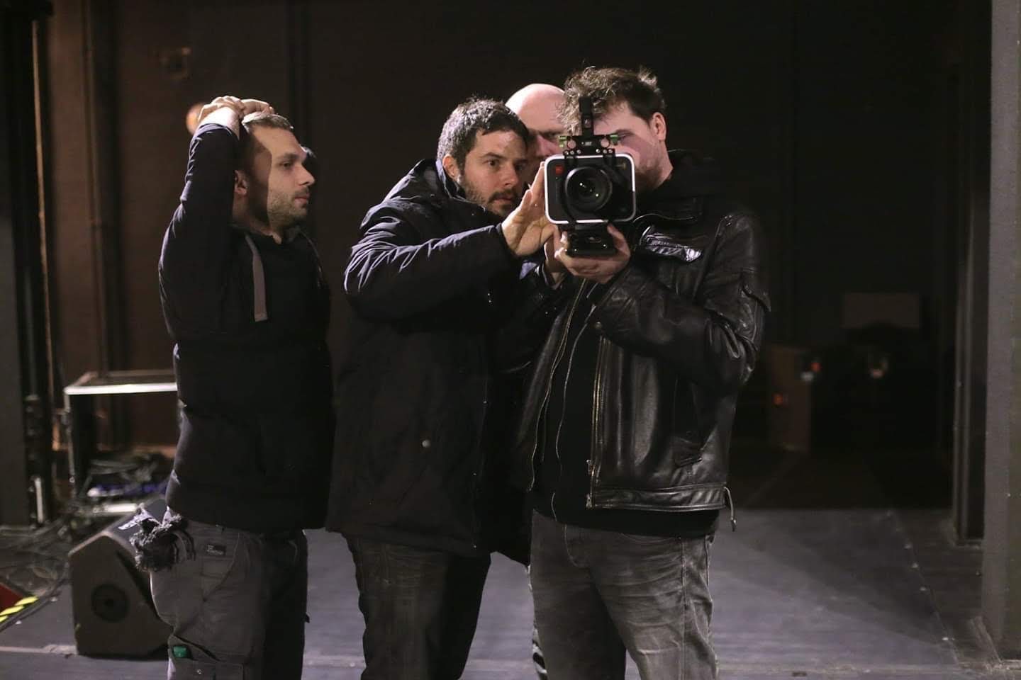Trzech mężczyzn na planie filmowym, jeden z nich trzyma kamerę, drugi pomaga mu ustawić ostrość, trzeci obserwuje.