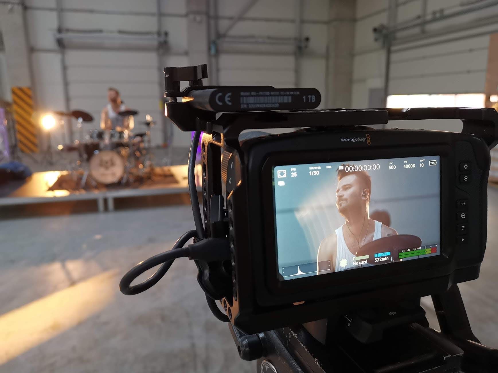 Ekran kamery Blackmagic Design podczas nagrywania perkusisty na scenie w hali, widoczny dysk 1TB podpięty do kamery.