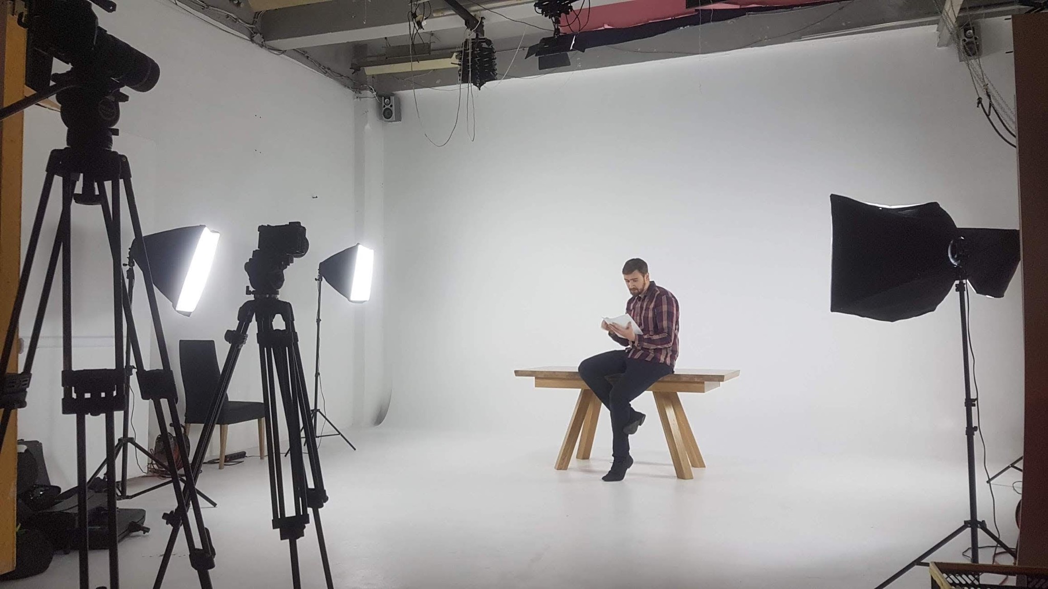 Studio filmowe z oświetleniem, kamerami na statywach i mężczyzną w koszuli w kratę siedzącym na drewnianym stole czytającym scenariusz.