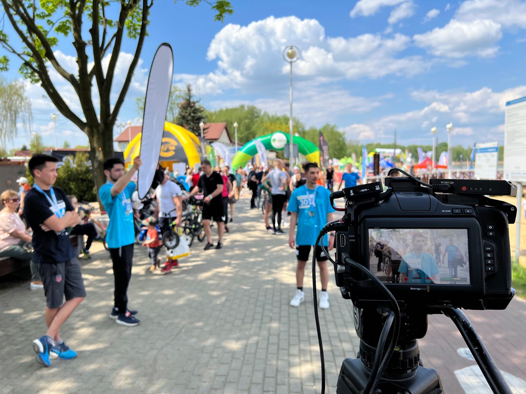Ekipa filmowa rejestruje przebieg imprezy sportowej. W kadrze widoczny profesjonalny aparat na statywie, dwóch asystentów z blendą oraz grupa uczestników biegu w tle.