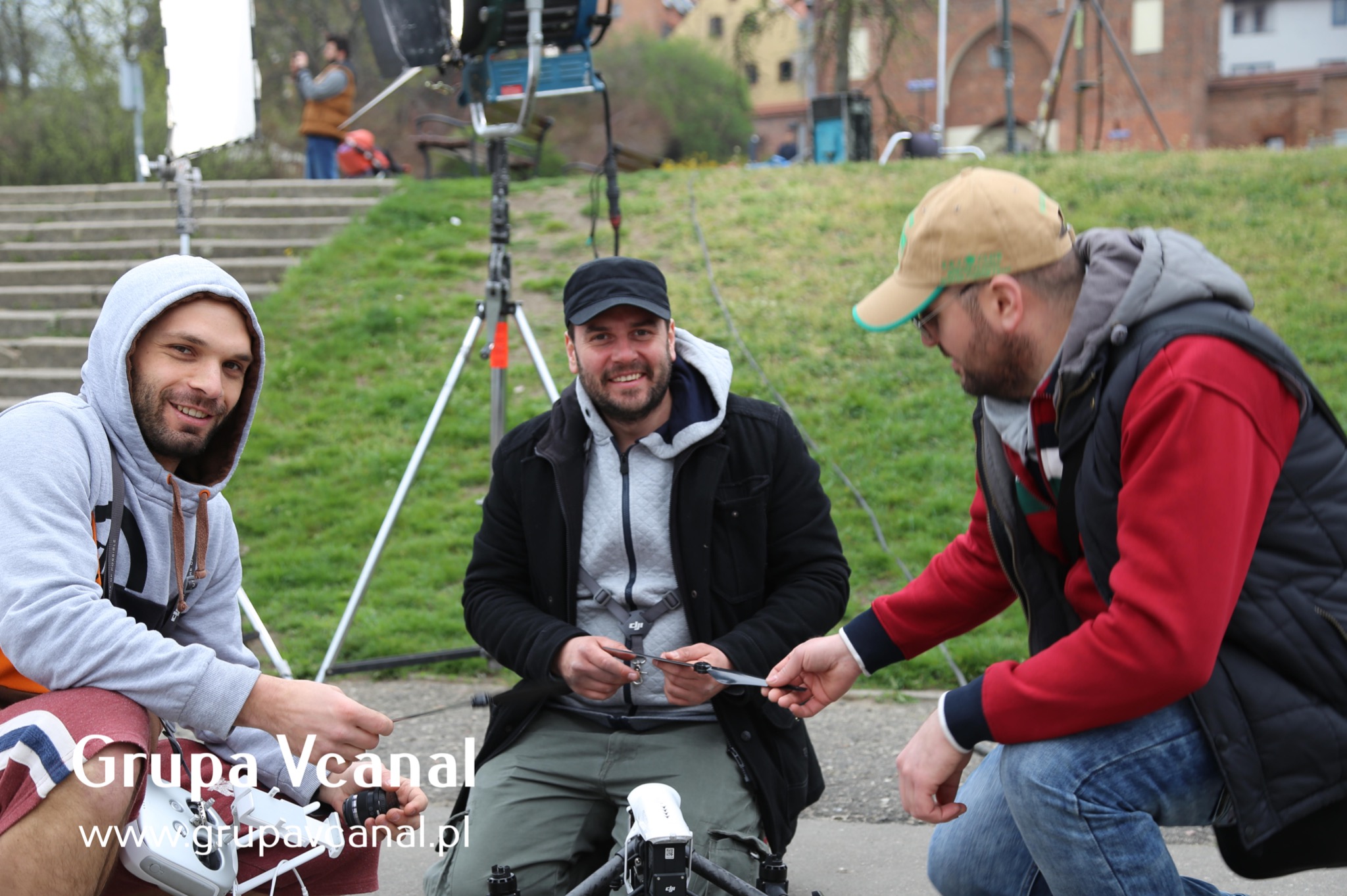 Trzech mężczyzn przygotowuje drona do lotu na tle schodów i zieleni, widoczny sprzęt oświetleniowy, jeden z mężczyzn trzyma śmigło drona.