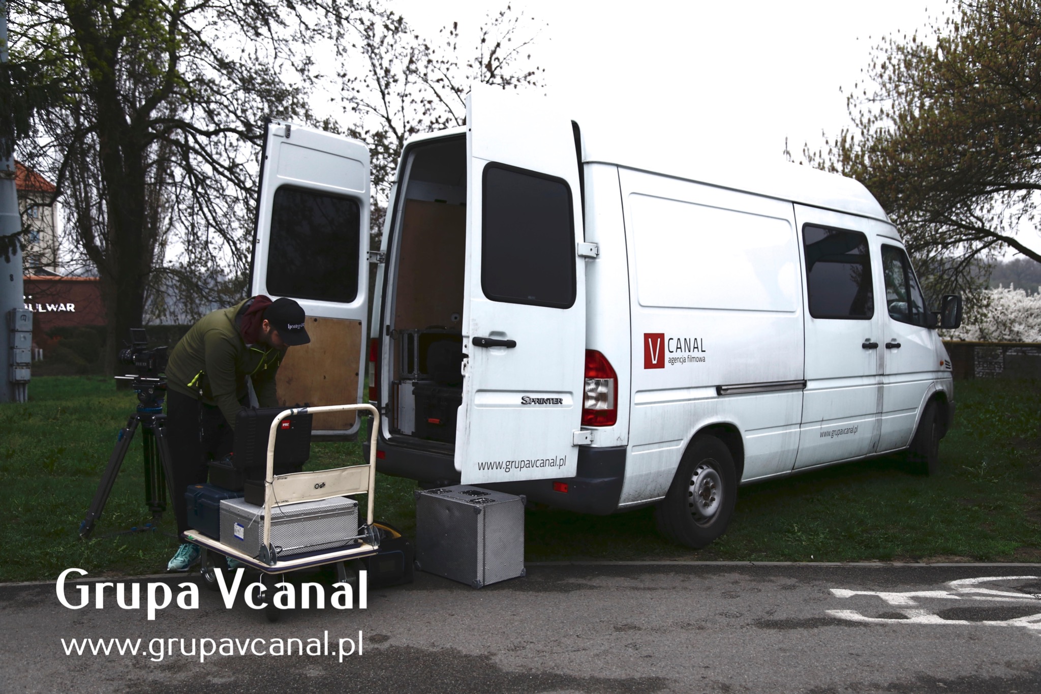 Ekipa filmowa rozładowuje sprzęt z białego vana z otwartymi drzwiami, w tle drzewa i budynek. Na wózku transportowym skrzynie ze sprzętem.