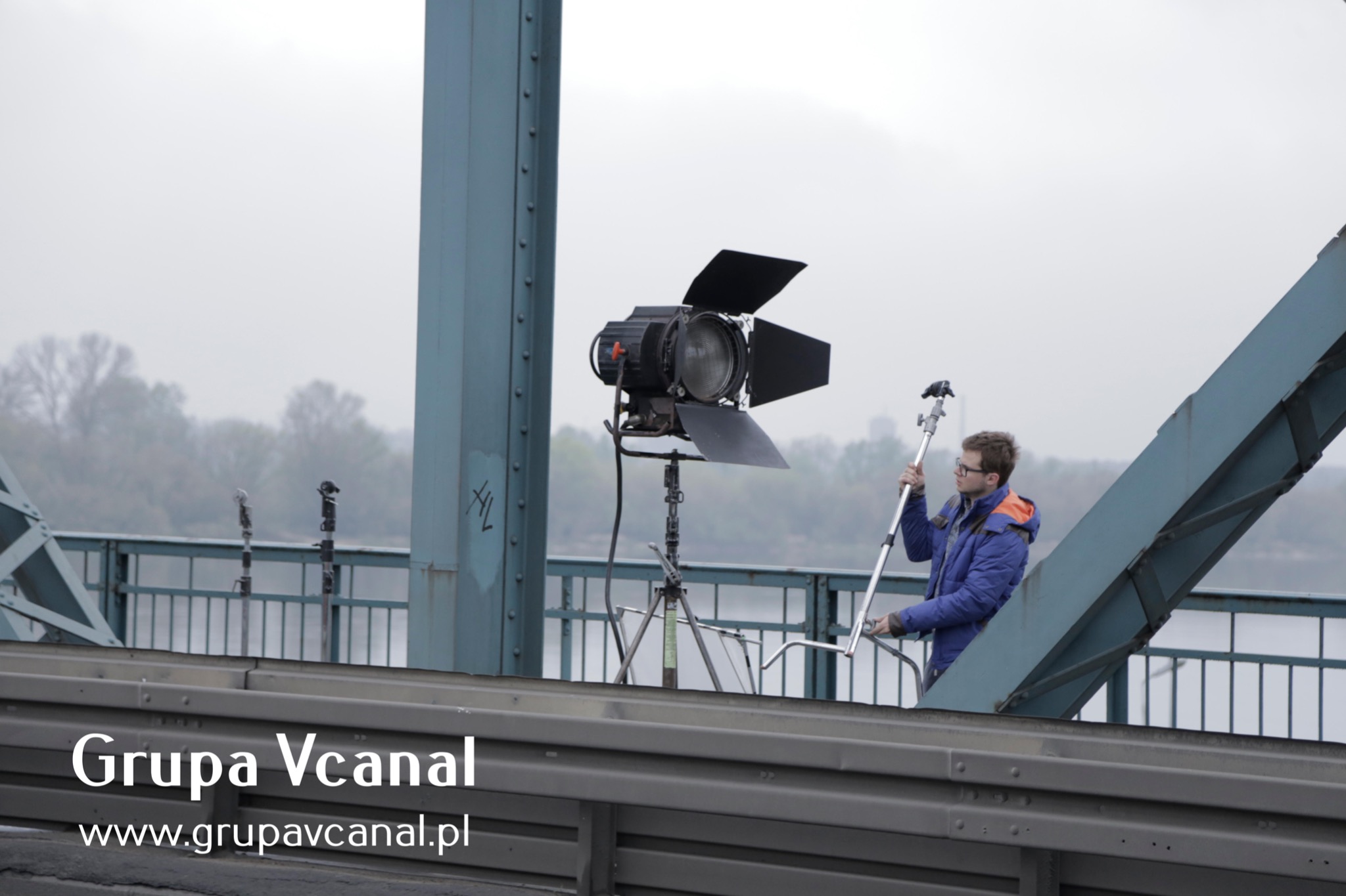 Ekipa filmowa ustawia oświetlenie na moście; widoczny reflektor studyjny z klapkami oraz mężczyzna w niebieskiej kurtce z pomarańczową podszewką trzymający statyw.
