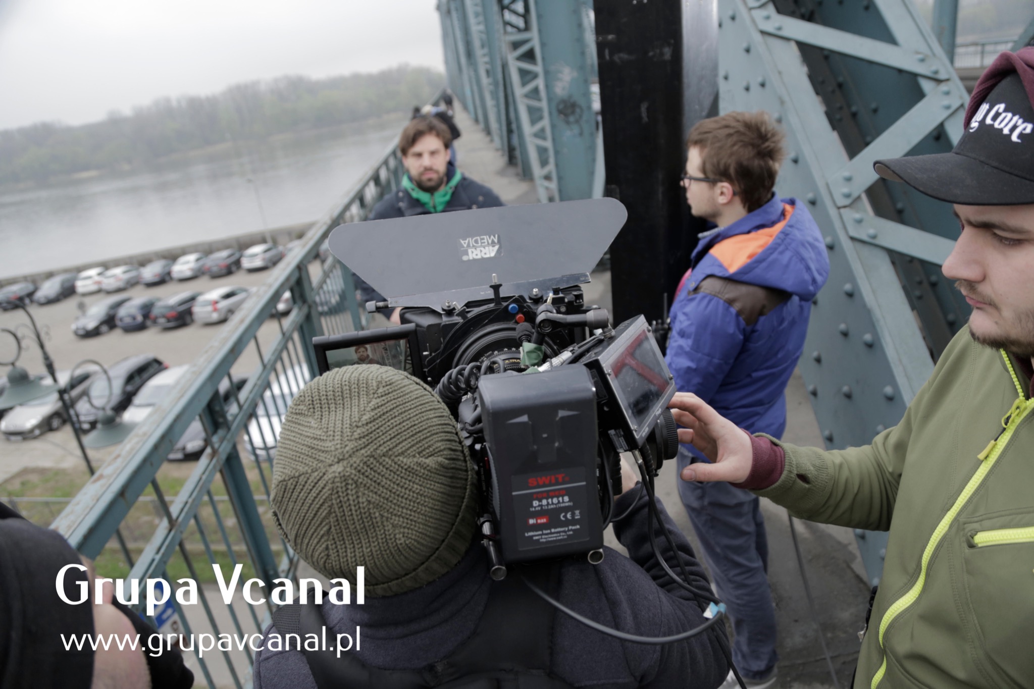 Ekipa filmowa kręci ujęcia na moście, widoczna profesjonalna kamera z osprzętem, w tle rzeka i zaparkowane samochody.