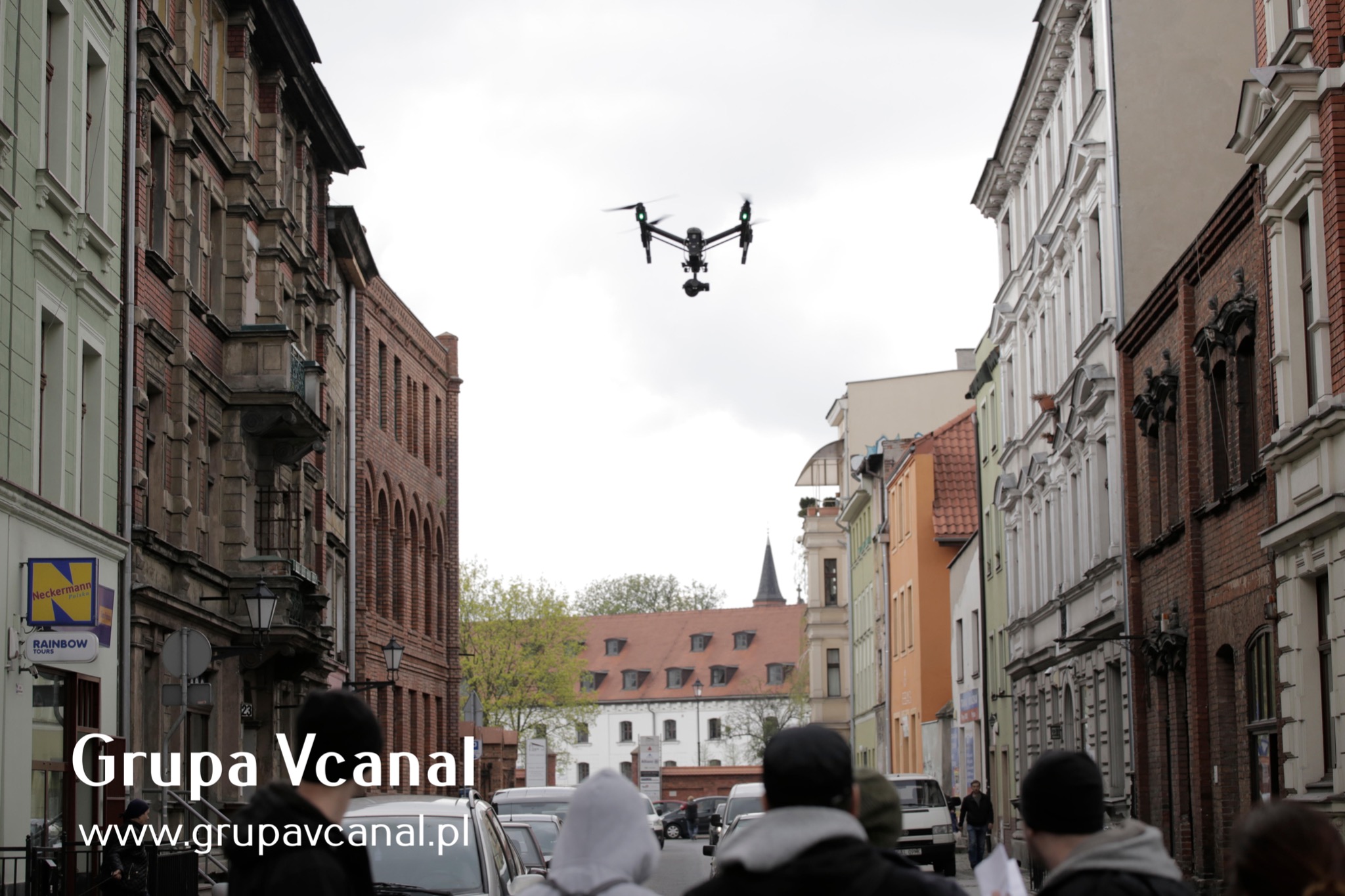 Czarny dron w locie nad ulicą miasta z zabudową kamieniczną, nagrywający materiał filmowy, widoczne sylwetki przechodniów i zaparkowane samochody