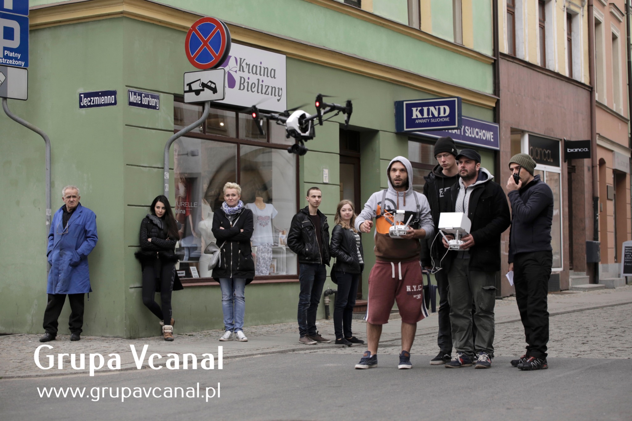 Ekipa filmowa z dronem na ulicy miejskiej podczas kręcenia ujęć, operator drona trzyma kontroler, w tle budynki z szyldami i przechodnie.