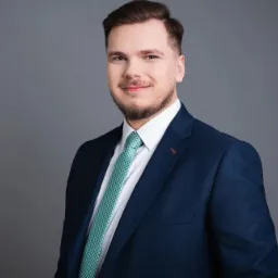 Młody mężczyzna w granatowym garniturze i zielonym krawacie, uśmiecha się na tle szarego studia. Profesjonalny portret.