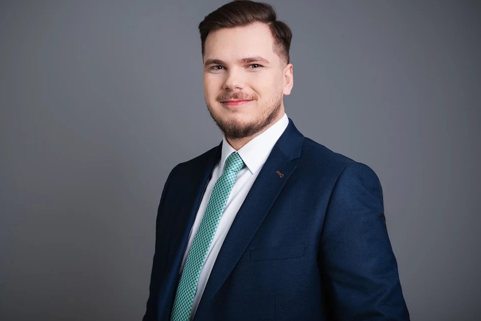 Młody mężczyzna w granatowym garniturze i zielonym krawacie, uśmiecha się na tle szarego studia. Profesjonalny portret.