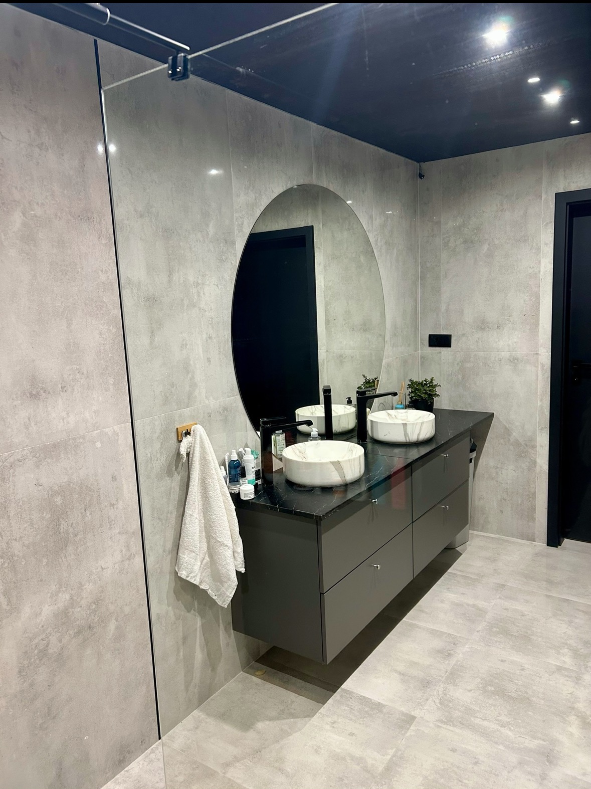 Nowoczesna łazienka z podwójną umywalką marmurkową na czarnym blacie, owalne lustro, szare płytki i kabina prysznicowa ze szkła. Minimalistyczny design.