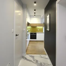 Wnętrze mieszkania: widok z korytarza na nowoczesną kuchnię z białymi szafkami, drewnianym blatem i żółtym panelem, marmurowa podłoga w korytarzu.
