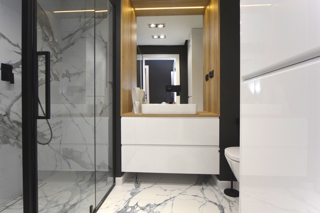 Nowoczesna łazienka z kabiną prysznicową wykończoną marmurowymi płytami, białą szafką z umywalką nablatową i podświetlanym lustrem w drewnianej obudowie.