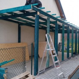 Krzysztof - Budowa zadaszenia tarasu z pomalowanymi na turkusowo drewnianymi słupami i belkami, widoczne płyty OSB, drabina aluminiowa i pracownik na górze konstrukcji.