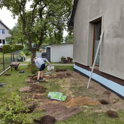 Krzysztof - Przygotowanie terenu pod wylewkę fundamentów: robotnik kopie łopatą wzdłuż ściany budynku ocieplonej styropianem, widoczne worki z betonem, wykopane dołki, sznurek wyznaczający linię, narzędzia...