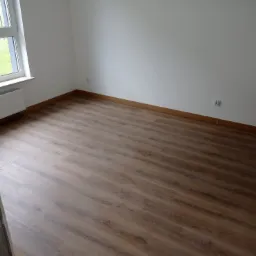 Puste pomieszczenie z brązowymi panelami podłogowymi, białymi ścianami i oknem z widokiem na zewnątrz. Listwa przypodłogowa w kolorze drewna. Widoczny biały grzejnik pod oknem.