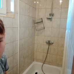 Krzysztof - Łazienka w trakcie remontu: widoczne uszkodzone płytki, odkręcona półka na kosmetyki, częściowo zdemontowana bateria prysznicowa, brudna wanna i narzędzia.