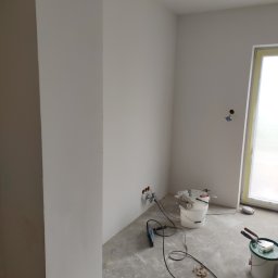Krzysztof - Pokój w trakcie prac wykończeniowych: świeżo pomalowane białe ściany, widoczne puszki elektryczne, narzędzia malarskie i wiaderka z farbą na podłodze pokrytej resztkami gładzi.