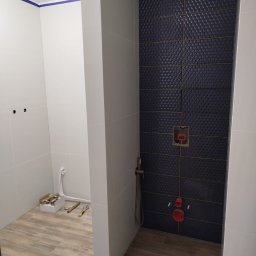 Krzysztof - Niedokończona łazienka z ciemnymi, strukturalnymi płytkami na ścianie prysznica, drewnopodobną podłogą i widocznymi instalacjami hydraulicznymi oraz narzędziami.